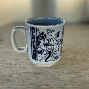 May include: A ceramic mug with a light blue exterior and a white interior. The mug features a dark blue floral design and vertical stripes. The handle is a matching light blue color. The mug is sitting on a wooden surface.