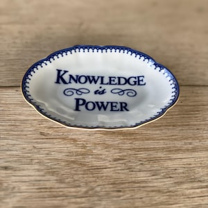 May include: Oval-shaped white ceramic dish with a blue scalloped edge and gold trim. The dish features the words "KNOWLEDGE is POWER" in blue script. The dish is on a wooden surface.