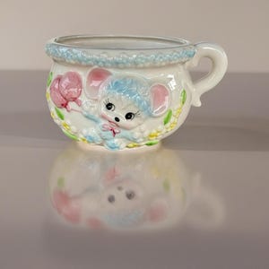 May include: A white ceramic baby cup with a handle, featuring a blue-haired mouse holding a pink balloon. The rim is decorated with a blue floral pattern, and the cup is adorned with yellow and green accents.