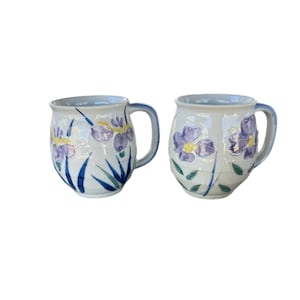 May include: Two ceramic mugs with a white base, adorned with hand-painted purple and yellow iris flowers and green leaves. The mugs have blue handles and rims, offering a touch of color and a floral design.