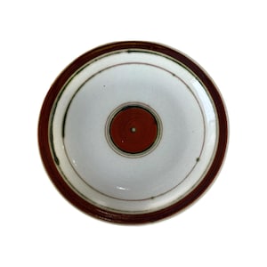 May include: A round ceramic plate with a white center and a dark brown rim. The center features a smaller, reddish-brown circle, with concentric rings of green and brown. The plate has a simple, yet elegant design.