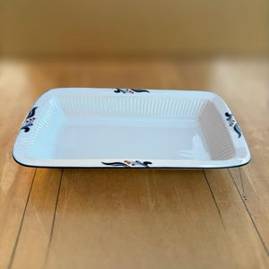 May include: A rectangular white ceramic baking dish with a ribbed interior surface. The rim features a dark blue and orange floral pattern. A portion of a measuring tape is visible, indicating a length of 15 inches along the side of the dish.