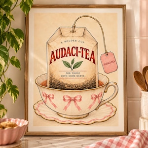 Op de afbeelding: Een ingelijste kunstdruk met een theezakje in een witte theekop met roze strikjes. Het theezakje is gelabeld met AUDACI-TEA, A BOLDER CUP, FOR THOSE WITH SOME NERVE. Een roze label aan het theezakje vermeldt ook AUDACI-TEA op een crèmekleurige achtergrond.
