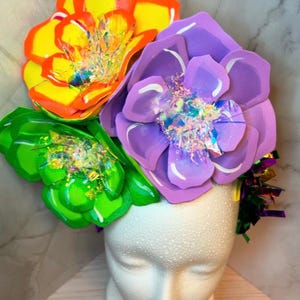 May include: A colorful flower crown with three large paper flowers in shades of yellow, green, and purple. The flowers have iridescent glitter accents and are arranged on a white mannequin head.
