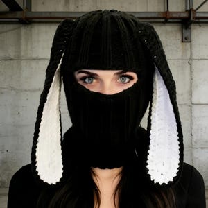 May include: Black knitted balaclava with long bunny ears. The ears are lined with white fabric. The face covering has an opening for the eyes. The person in the image has blue eyes and dark hair.