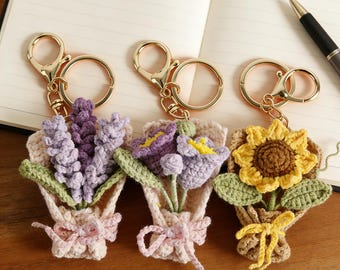 May include: Three crochet flower bouquet keychains. The bouquets feature lavender, purple flowers, and a sunflower. Each bouquet is wrapped in a beige paper-like material and tied with a bow. The keychains have gold-colored clasps.