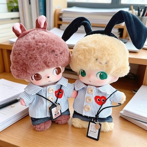 May include: Two plush dolls with large eyes and fluffy hair, one in a brown bunny hat and the other in a black bunny hat. Both dolls wear striped shirts with heart patches and denim shorts. Each doll has a small tag around its neck.