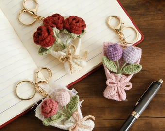 May include: Three crochet flower bouquet keychains. One has red roses, another has pink and peach flowers, and the third has purple and lavender flowers. Each bouquet is tied with a bow and attached to a gold keychain.