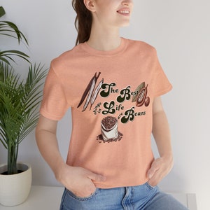 May include: A peach colored t-shirt with a graphic print of beans, cocoa pods, and vanilla beans. The text on the shirt reads "The Best Things in Life are Beans".