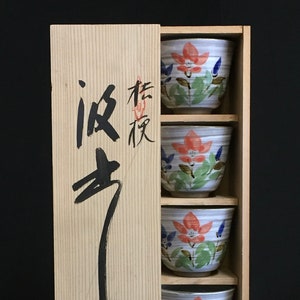 以下が含まれることがあります： ピンク、ブルー、グリーンの色合いの花柄が描かれた、白い小さな陶器のボウルが5つセットになっています。ボウルは、前面に日本語の書道が書かれた木製の箱の中に積み重ねられています。