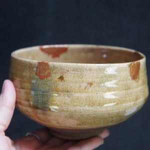 May include: A beige ceramic bowl with a textured surface and a wide, rounded shape. The bowl has brown and red accents and white horizontal lines. The bowl is held in a hand against a dark background. The bowl is approximately 13 cm in diameter.