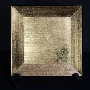以下が含まれることがあります： 正方形の金色の装飾プレート。表面はテクスチャ加工が施され、緑色の針とベリーの小枝が描かれています。プレートには隆起したテクスチャ加工の縁があり、黒いスタンドに展示されています。