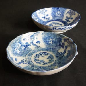 以下が含まれることがあります： 2つのアンティークの青と白の磁器のボウル。ボウルは、複雑な花柄と幾何学模様が特徴です。縁はスカラップエッジです。ボウルは暗い背景に配置されています。