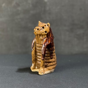 May include: A small ceramic figurine of a brown and tan mythical creature with a textured surface. The creature has a wide mouth with small holes and a raised brow.
