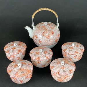 May include: A white teapot with a brown wicker handle and a floral design. The teapot is surrounded by six white teacups with floral designs.