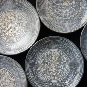 May include: Overhead shot of five gray ceramic plates with a floral pattern in the center. The plates have a textured rim and a dark gray color. The floral pattern is white and the plates are arranged on a black surface.
