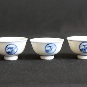 以下が含まれることがあります： 青と白の模様が施された、小さな白い陶磁器のティーカップが5つ。各カップは小さな足と丸い形をしています。