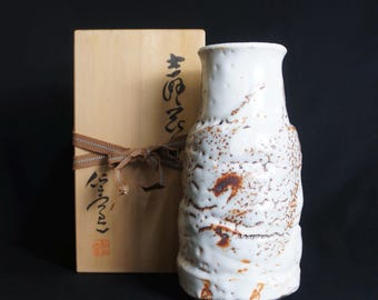 May include: A white ceramic vase with a textured surface and brown speckles. The vase is in a wooden box with Japanese writing on it.