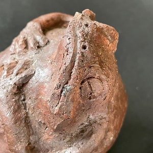 Peut inclure: Une sculpture en céramique brune d'une grenouille avec les bras croisés devant elle. La grenouille a une surface texturée et un visage détaillé.