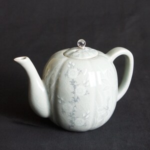 以下が含まれることがあります： 蓋に透明なガラスのノブが付いた、薄い緑色のセラミック製のティーポット。花柄のデザインが施されています。