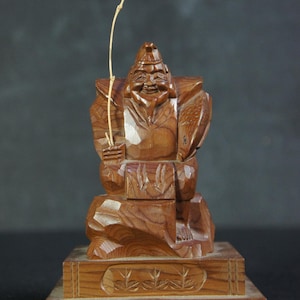May include: A detailed wooden carving of a smiling figure holding a fishing rod, seated on a carved base. The figure has a hat and a fish-shaped shield. The base has a decorative carved design. The wood has a warm, natural tone.