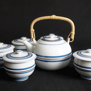 Puede incluir: Una tetera blanca con asa de bambú y rayas azules y marrones, junto con cinco tazas de té y platillos a juego.