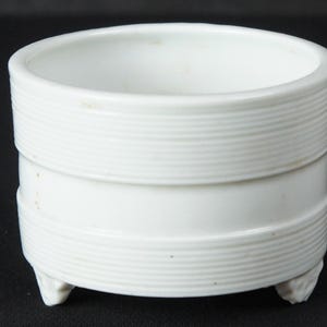 May include: A white ceramic planter with a cylindrical shape and three small feet. The planter features horizontal ribbed detailing around the middle and top sections. The interior is smooth and open, suitable for small plants or decorative items.