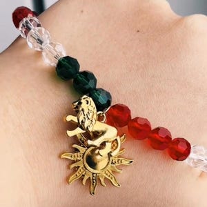 May include: A bracelet featuring red, green, and clear faceted beads. A gold-coloured charm shows a lion over a sunburst. The bracelet is a fashion accessory.