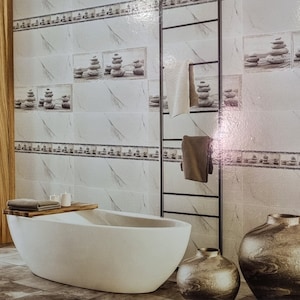 Può includere: Un bagno con piastrelle bianche effetto marmo con opere d'arte di pietre impilate. Una vasca ovale bianca, dettagli in legno e vasi decorativi completano l'ambiente simile a una spa. Un portasciugamani nero.