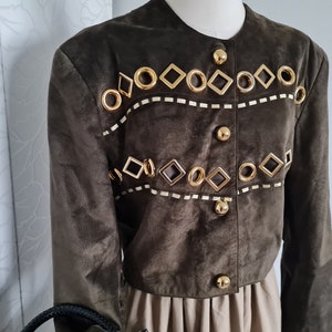 Può includere: Una giacca di camoscio marrone con decorazioni e bottoni dorati. La giacca ha un collo rotondo e maniche lunghe.