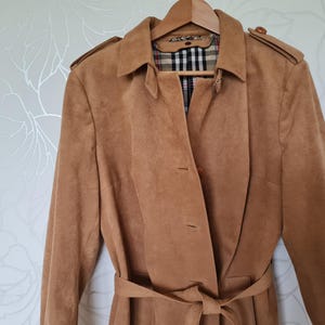 Könnte beinhalten: Ein hellbrauner Trenchcoat mit Gürtel, Knöpfen und Schulterklappen. Der Mantel hängt an einem Holzbügel. Das Innenfutter hat ein Karomuster.