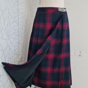 Può includere: Gonna a portafoglio con motivo a quadri rossi e blu navy. La gonna ha un bordo sfrangiato e una fibbia color argento. Il tessuto è di peso medio, adatto alle temperature più fresche. La gonna è esposta su un manichino.