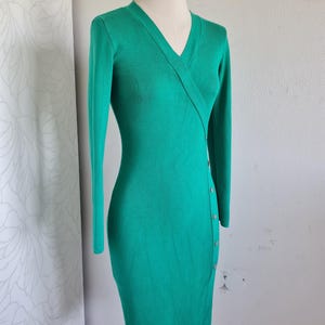 Könnte beinhalten: Smaragdgrünes, langärmliges Kleid mit V-Ausschnitt und Wickeldesign. Das Kleid hat Knöpfe an der Seite und wird auf einer Schaufensterpuppe präsentiert. Das Kleid ist aus Strickstoff gefertigt, ca. 100 cm lang.