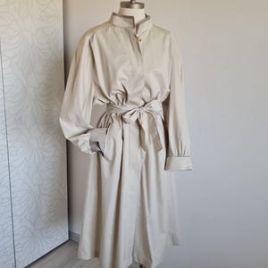 May include: A beige trench coat with a mandarin collar, long sleeves, and a matching belt tied at the waist. The coat has a button closure and pockets. The coat is displayed on a mannequin.