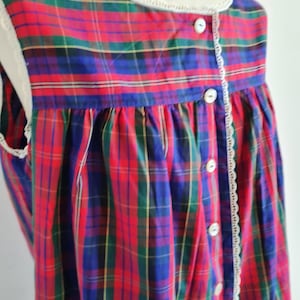 Puede incluir: Camisón sin mangas de estilo vintage con un vibrante estampado de cuadros rojos, azules, verdes y amarillos. El camisón presenta un ribete de encaje blanco a lo largo del escote y las sisas, y una fila de botones blancos en la parte delantera.