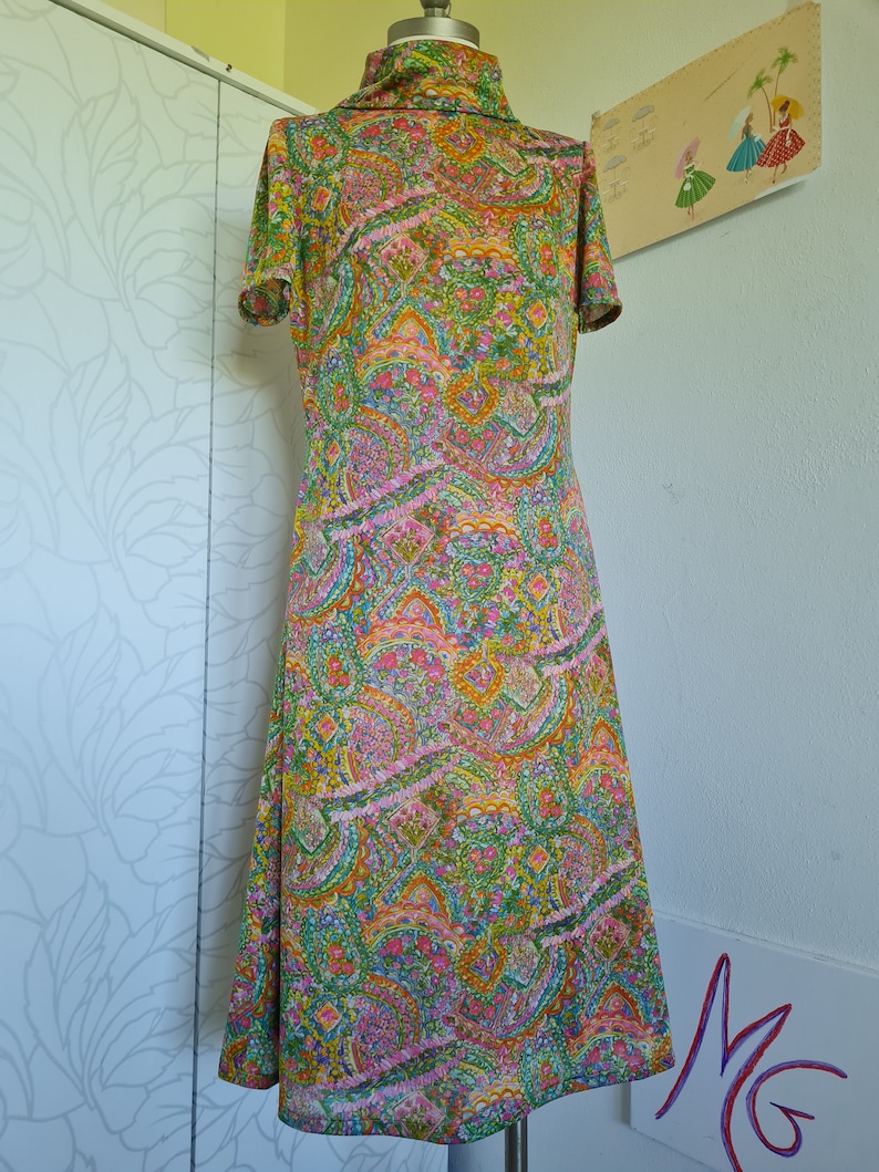 Pu&ograve; includere: Un abito vintage con stampa paisley in tonalit&agrave; di verde, rosa, arancio e giallo. L'abito ha un collo alto e maniche corte.