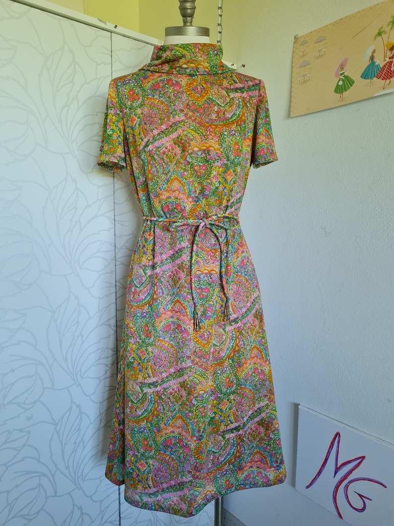 Pu&ograve; includere: Un abito vintage con stampa paisley in tonalit&agrave; di verde, rosa, arancio e giallo. L'abito ha un collo alto, maniche corte e una cintura con lacci.