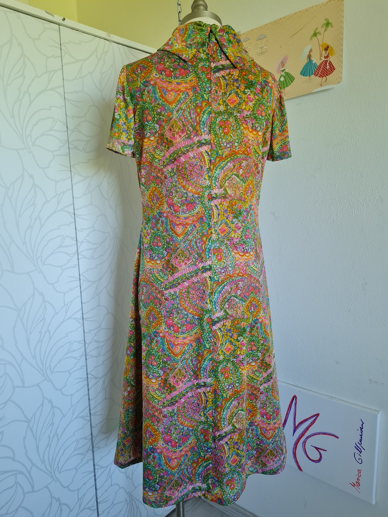 Pu&ograve; includere: Un abito vintage con stampa paisley in tonalit&agrave; di verde, rosa, arancio e giallo. L'abito ha un colletto alto e maniche corte.
