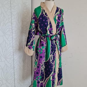 Op de afbeelding: Een kleurrijke badjas met een nautisch ontwerp. De badjas heeft een patroon van paarse stippen, groene panelen en witte touwaccenten. De kraag en manchetten zijn beige en de badjas wordt in de taille vastgebonden met een bijpassende riem.