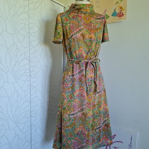Pu&ograve; includere: Un abito vintage con stampa paisley in tonalit&agrave; di verde, rosa e giallo. L'abito ha un collo alto, maniche corte e una cintura con laccio in vita.