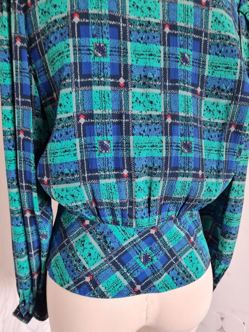 May include: A blue and green plaid blouse with a gathered waist. The blouse has long sleeves and a button-up front.