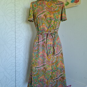Pu&ograve; includere: Un abito vintage degli anni '70 con stampa paisley in tonalit&agrave; di verde, rosa, arancio e giallo. L'abito ha un collo alto, maniche corte e una cintura con lacci.