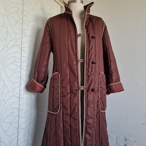 May include: A long, quilted, rust-brown coat featuring vertical stitching, a high collar, and three toggle closures down the front. The garment includes two large patch pockets on the front, each finished with light-colored piping along the edges.