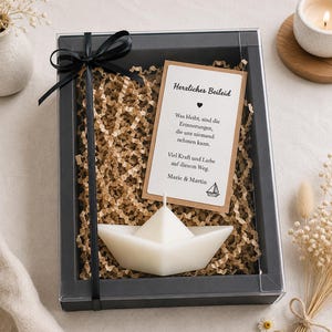 Puede incluir: Una caja de regalo negra que contiene una vela blanca con forma de barco de papel, colocada sobre relleno de papel marrón triturado. Se incluye una tarjeta rectangular con el texto en alemán Herzliches Beileid, Was bleibt, sind die Erinnerungen, die uns niemand nehmen kann, Viel Kraft und Liebe auf diesem Weg, Marie & Martin. La caja está atada con una cinta negra.