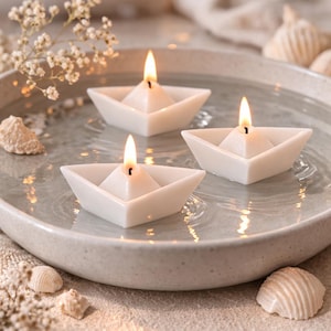 Peut inclure: Trois bougies blanches en forme de bateau avec des flammes allumées flottent dans un bol peu profond rempli d'eau. Les bougies sont entourées de petits coquillages et de délicates fleurs blanches, créant un thème serein et nautique.