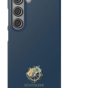 Puede incluir: Una funda de smartphone azul oscuro con un recorte vertical para triple c&aacute;mara y un gr&aacute;fico circular de un tim&oacute;n de barco adornado con flores claras y hojas verdes. El texto K&Uuml;STENLIEBE est&aacute; impreso en color dorado debajo del tim&oacute;n floral.
