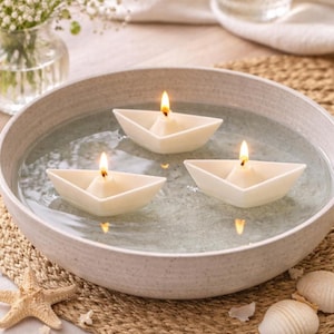May include: A serene display of three lit, white paper boat candles floating in a water-filled, speckled ceramic bowl. The bowl rests on a woven mat, with a starfish and seashells. A vase of white flowers and a glass are in the background.