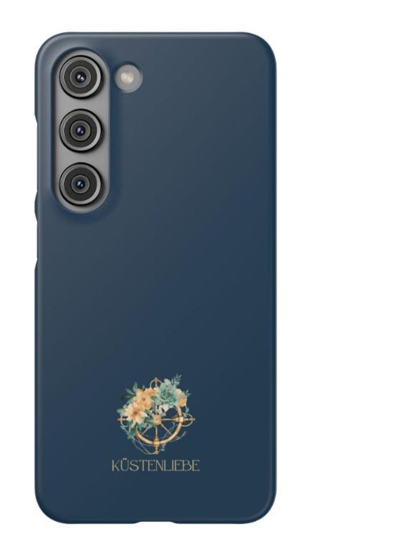 May include: A dark blue smartphone case featuring a vertical triple camera cutout and a circular graphic of a ship's wheel adorned with light-colored flowers and green leaves. The text K&Uuml;STENLIEBE is printed in gold below the floral ship's wheel.