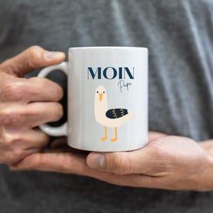 May include: White ceramic mug with a blue and black seagull illustration and the text "Moin Papa" in blue.