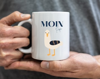 Puede incluir: Taza de cerámica blanca con una ilustración de gaviota azul y negra y el texto "Moin Papa" en azul.